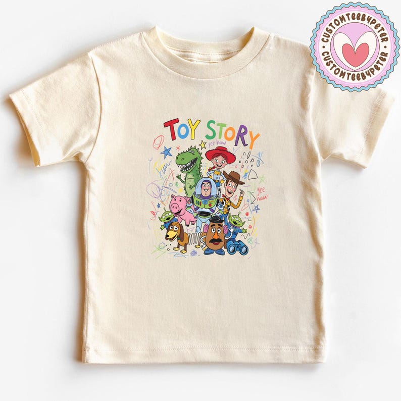 May include: A cream-colored t-shirt featuring a colorful graphic of characters from the animated film "Toy Story." The text "TOY STORY" is written above the characters. The shirt has short sleeves and a crew neck.