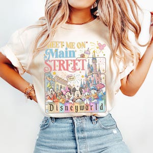 May include: Cream-colored t-shirt featuring the text "Meet Me On Main Street" above a graphic of Disney characters and Cinderella's castle. The graphic includes Mickey Mouse, Minnie Mouse, Donald Duck, and other characters. The word "Disneyworld" is at the bottom.