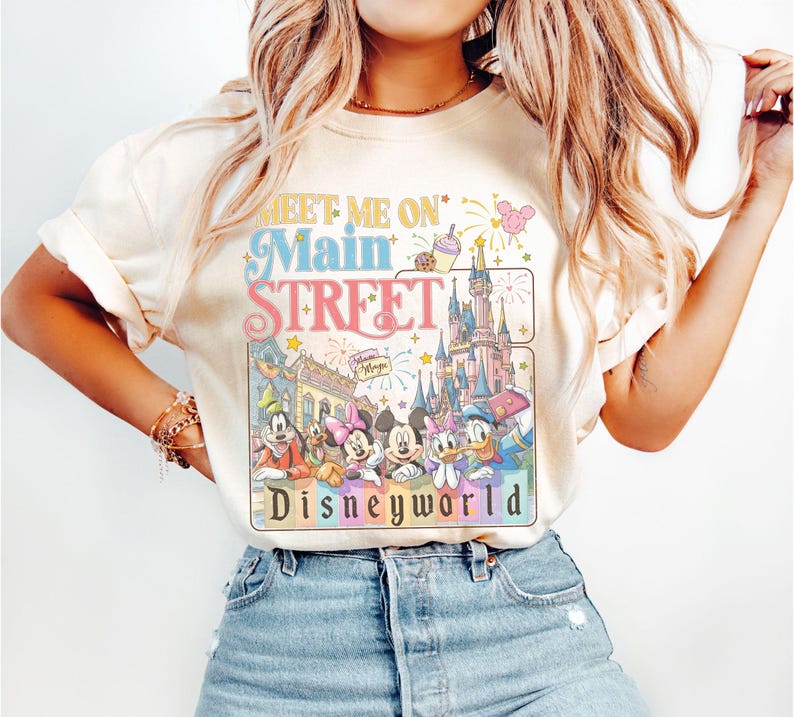 May include: Cream-colored t-shirt featuring the text "Meet Me On Main Street" above a graphic of Disney characters and Cinderella's castle. The graphic includes Mickey Mouse, Minnie Mouse, Donald Duck, and other characters. The word "Disneyworld" is at the bottom.