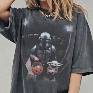 May include: Dark gray t-shirt featuring a graphic of The Mandalorian and Grogu, with a jack-o'-lantern candy bucket. The image is set against a nighttime suburban backdrop. The shirt has a vintage, worn look.