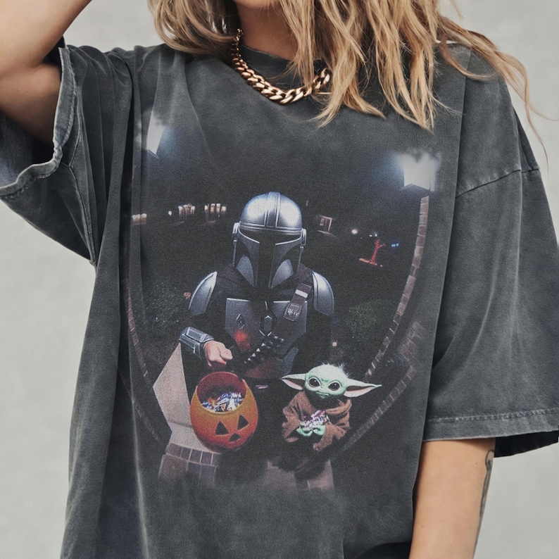 May include: Dark gray t-shirt featuring a graphic of The Mandalorian and Grogu, with a jack-o'-lantern candy bucket. The image is set against a nighttime suburban backdrop. The shirt has a vintage, worn look.
