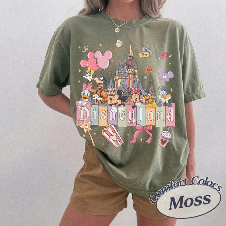 May include: Olive green t-shirt featuring a Disneyland graphic with Mickey Mouse, Minnie Mouse, Donald Duck, and other Disney characters. The shirt has the words "Disneyland" and "Comfort Colors Moss" printed on it.