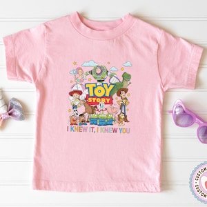 May include: A light pink short-sleeve t-shirt featuring a colorful graphic of Toy Story characters including Woody, Buzz Lightyear, Jessie, and others. The text "TOY STORY" is printed in the center, with "I KNEW IT, I KNEW YOU" below the characters.