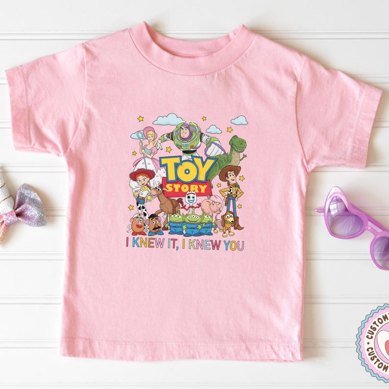 May include: A light pink short-sleeve t-shirt featuring a colorful graphic of Toy Story characters including Woody, Buzz Lightyear, Jessie, and others. The text "TOY STORY" is printed in the center, with "I KNEW IT, I KNEW YOU" below the characters.