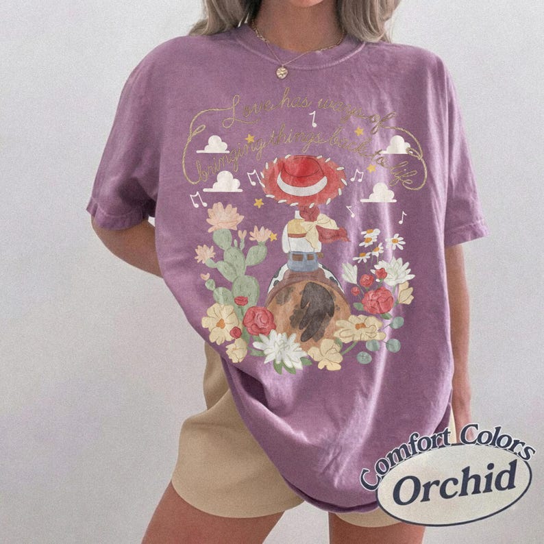 May include: A purple short-sleeve t-shirt featuring a graphic of a cowgirl figure on a horse, surrounded by cacti, flowers, clouds, and musical notes. The text reads Love has a way of bringing things back to life in a cursive gold script font.