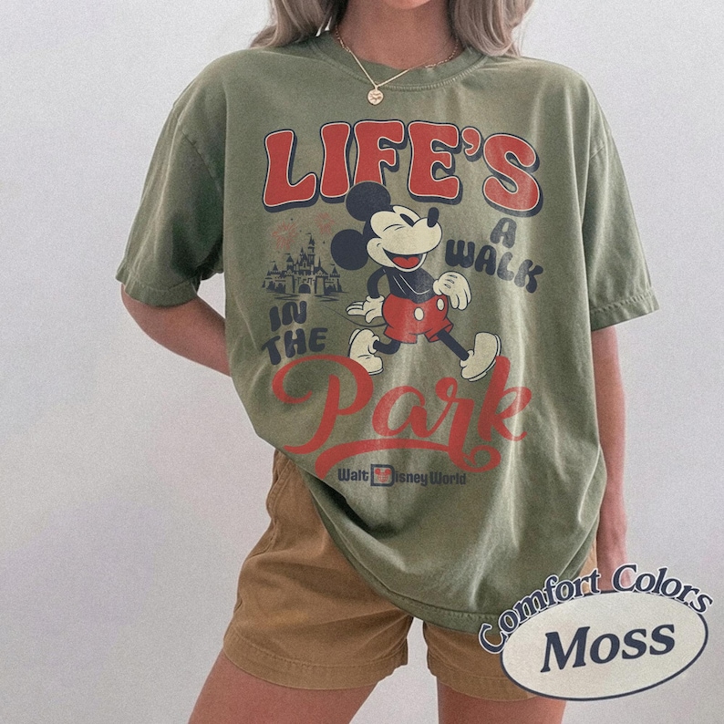 May include: An olive green t-shirt with a graphic of Mickey Mouse and the text "LIFE'S A WALK IN THE PARK" in red. The shirt also includes the words "Walt Disney World". The shirt is labelled "Comfort Colors Moss".