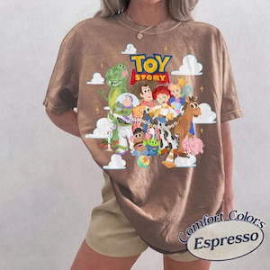 May include: A brown short-sleeve t-shirt featuring a graphic print of Toy Story characters including Woody, Buzz Lightyear, Rex, Jessie, and others. The design includes white clouds, yellow stars, and the Toy Story logo. A label reads Comfort Colors Espresso.