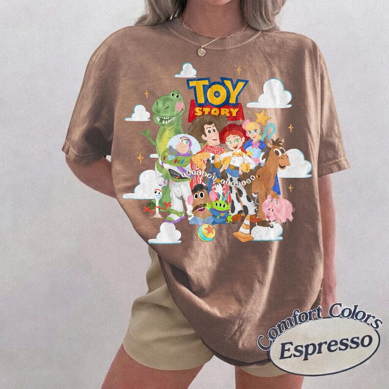 May include: A brown short-sleeve t-shirt featuring a graphic print of Toy Story characters including Woody, Buzz Lightyear, Rex, Jessie, and others. The design includes white clouds, yellow stars, and the Toy Story logo. A label reads Comfort Colors Espresso.