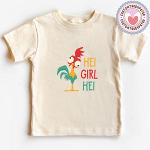 Peut inclure: Un t-shirt cr&egrave;me avec un coq de dessin anim&eacute; et le texte "HEI GIRL HEI" en jaune, rouge et vert. Le coq a des d&eacute;tails rouges, verts, jaunes et blancs. Un logo circulaire avec un c&oelig;ur est dans le coin sup&eacute;rieur droit.