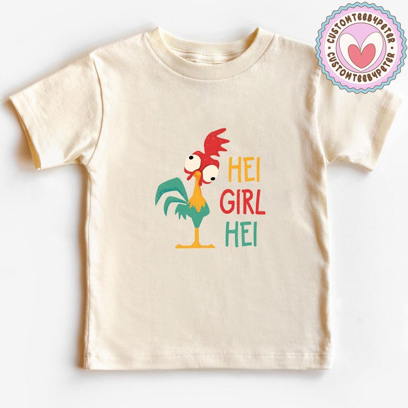Peut inclure: Un t-shirt cr&egrave;me avec un coq de dessin anim&eacute; et le texte "HEI GIRL HEI" en jaune, rouge et vert. Le coq a des d&eacute;tails rouges, verts, jaunes et blancs. Un logo circulaire avec un c&oelig;ur est dans le coin sup&eacute;rieur droit.