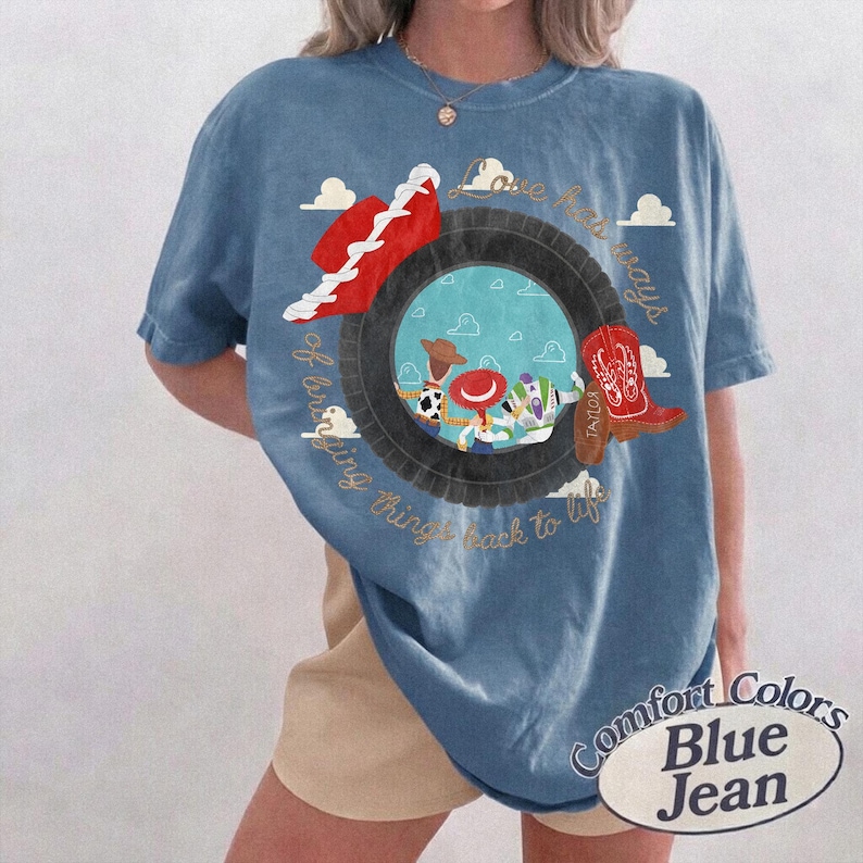 May include: A blue t-shirt featuring a graphic of a black tire framing a scene with cartoon characters, a red cowboy hat, and a red cowboy boot. The text reads Love has ways of bringing things back to life. The shirt has a crew neck and short sleeves.
