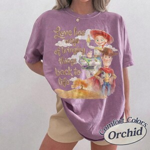 K&ouml;nnte beinhalten: Ein violettes Kurzarm-T-Shirt mit einer Grafik von Woody, Buzz Lightyear und Jessie aus Toy Story. Der Text lautet, Love has ways of bringing things back to life. Das Shirt hat einen Rundhalsausschnitt, eine lockere Passform und ein Etikett.