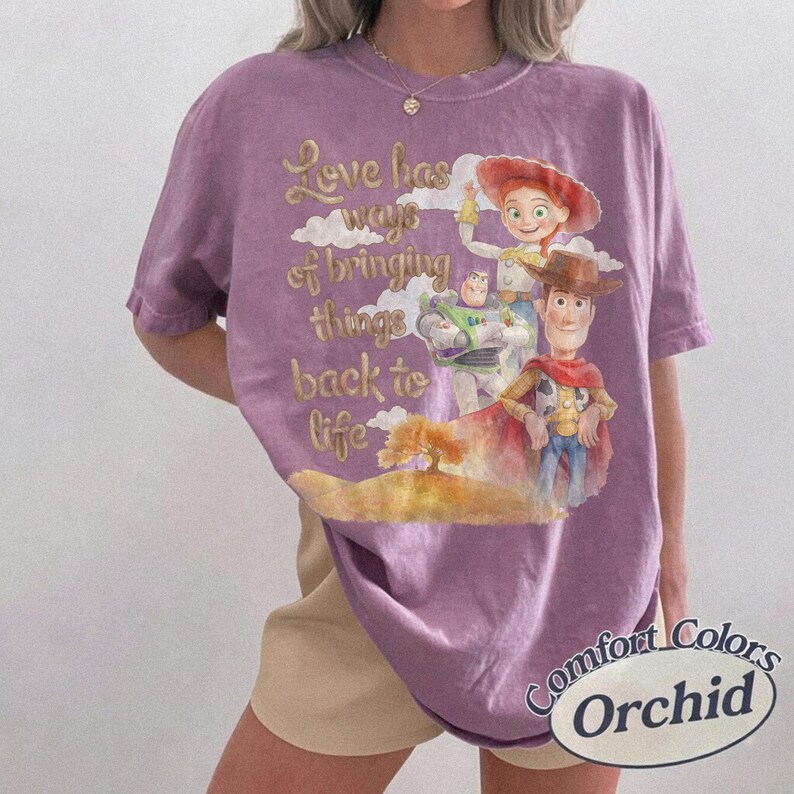K&ouml;nnte beinhalten: Ein violettes Kurzarm-T-Shirt mit einer Grafik von Woody, Buzz Lightyear und Jessie aus Toy Story. Der Text lautet, Love has ways of bringing things back to life. Das Shirt hat einen Rundhalsausschnitt, eine lockere Passform und ein Etikett.