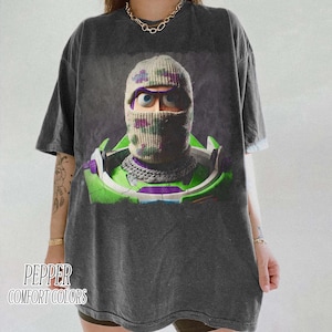 May include: A charcoal gray short-sleeve t-shirt featuring a graphic print of a character wearing a camouflage balaclava over their head. The character has large blue eyes and is wearing a green and white space-themed suit with a silver chain.