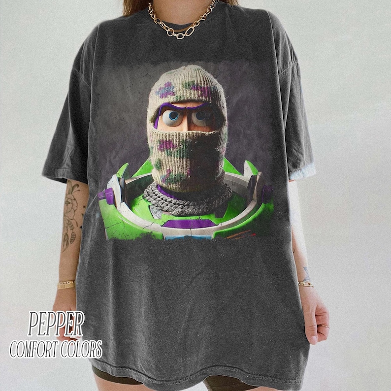 May include: A charcoal gray short-sleeve t-shirt featuring a graphic print of a character wearing a camouflage balaclava over their head. The character has large blue eyes and is wearing a green and white space-themed suit with a silver chain.