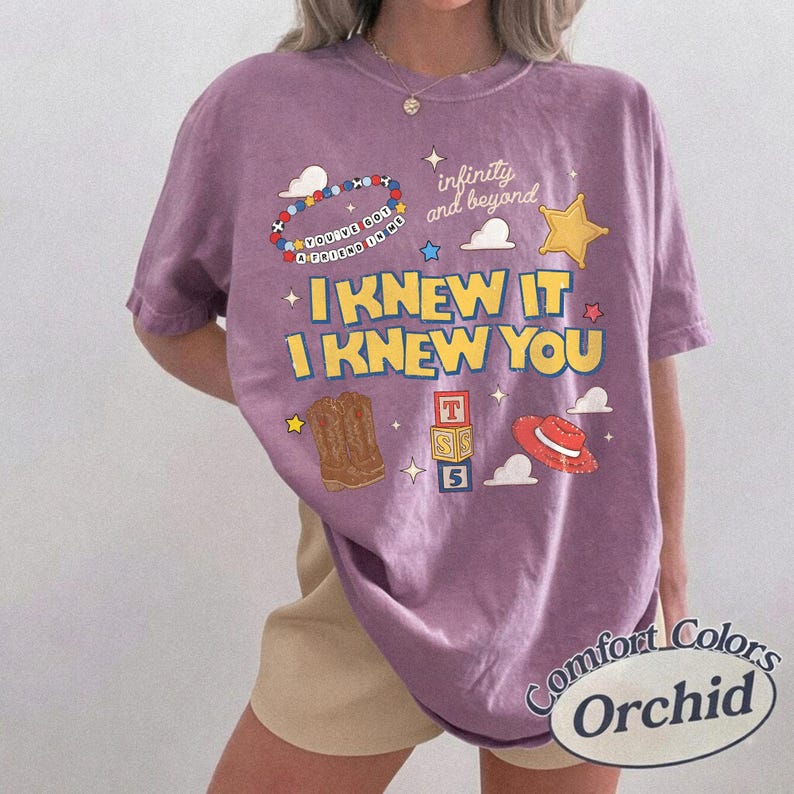 May include: A purple short-sleeve t-shirt featuring a graphic print with the text "I KNEW IT I KNEW YOU", "infinity and beyond", and "YOU'VE GOT A FRIEND IN ME". The design includes a beaded bracelet, a yellow star, cowboy boots, blocks, and a hat.