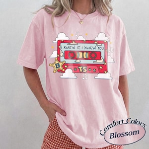 May include: A light pink t-shirt featuring a graphic of a red cassette tape with the text I KNEW IT, I KNEW YOU and TS. The design includes white clouds, yellow stars, and musical notes. The shirt has a crew neckline and short sleeves.