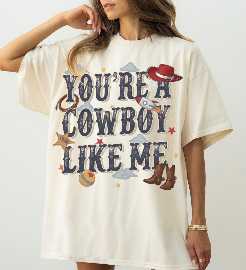 May include: An off-white short-sleeve t-shirt featuring the dark blue text "YOU'RE A COWBOY LIKE ME" surrounded by graphic illustrations of a red cowboy hat, a rocket, a lasso, a sheriff star, a colorful ball, brown cowboy boots, and small stars.