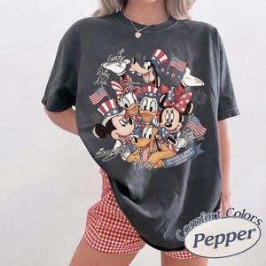 Puede incluir: Camiseta Comfort Colors gris oscuro con un dise&ntilde;o patri&oacute;tico de Disney. Presenta a Mickey Mouse, Minnie Mouse, Donald Duck, Daisy Duck, Goofy y Pluto, junto con banderas americanas y el texto "America 250th USA Anniversary 1776-2026."