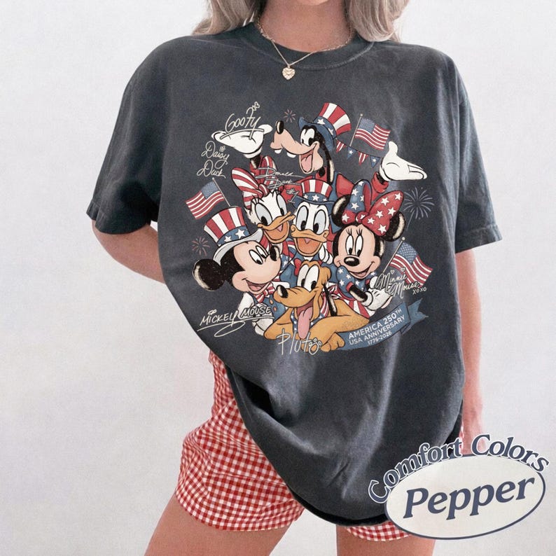 Puede incluir: Camiseta Comfort Colors gris oscuro con un dise&ntilde;o patri&oacute;tico de Disney. Presenta a Mickey Mouse, Minnie Mouse, Donald Duck, Daisy Duck, Goofy y Pluto, junto con banderas americanas y el texto "America 250th USA Anniversary 1776-2026."