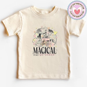 May include: A cream-coloured t-shirt with a graphic design. The design features the word "MAGICAL" and "PLACE ON EARTH" with illustrations of a castle and characters in black, pink, and yellow. A circular logo is visible on the upper right of the t-shirt.