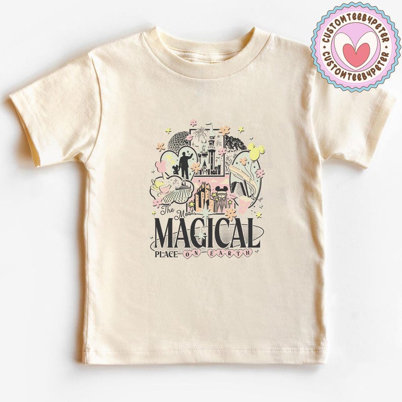 May include: A cream-coloured t-shirt with a graphic design. The design features the word "MAGICAL" and "PLACE ON EARTH" with illustrations of a castle and characters in black, pink, and yellow. A circular logo is visible on the upper right of the t-shirt.