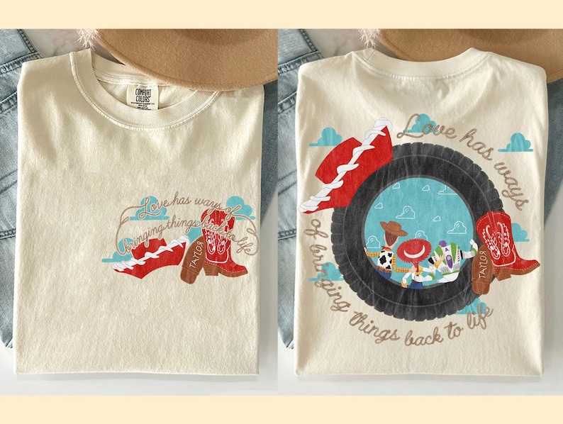 May include: A cream-coloured t-shirt featuring a graphic print of a tyre, cowboy boots, a red hat, and cartoon characters. The text reads, Love has ways of bringing things back to life. The design appears on the front chest and in a larger version on the back.