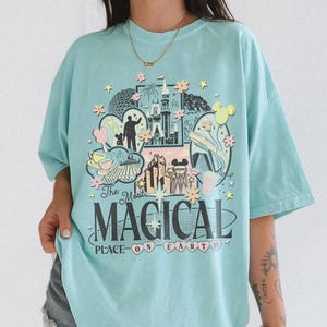 May include: A pale blue t-shirt with a vibrant graphic design depicting a castle, fireworks, and Mickey Mouse ears. The text "The Most MAGICAL PLACE ON EARTH" is printed on the front. The shirt has short sleeves and a loose fit.