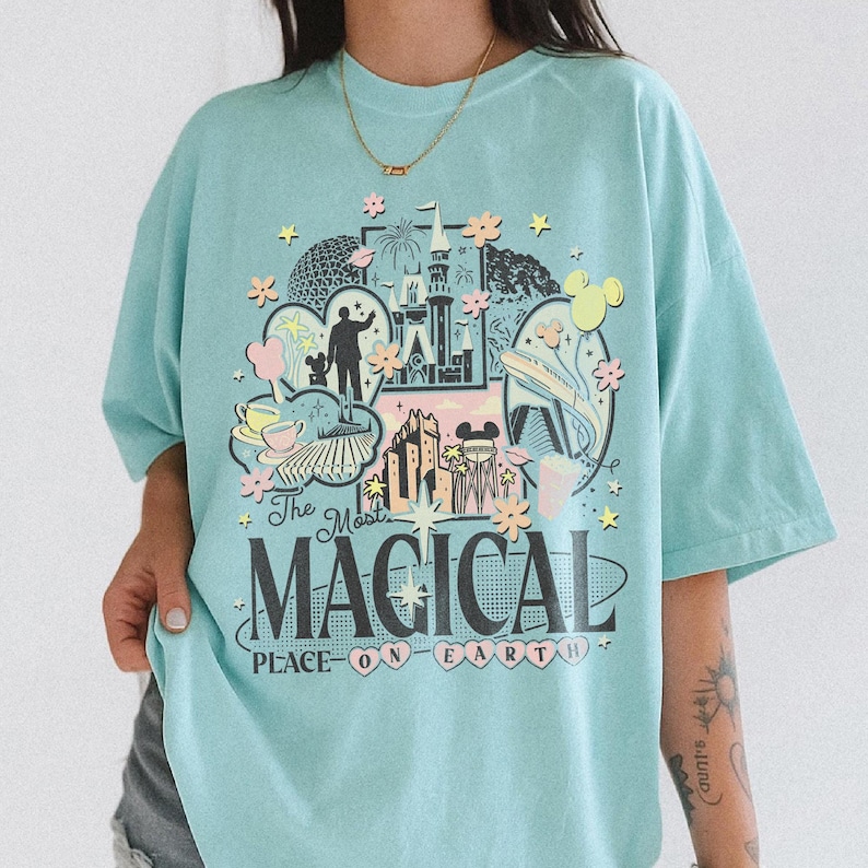 May include: A pale blue t-shirt with a vibrant graphic design depicting a castle, fireworks, and Mickey Mouse ears. The text "The Most MAGICAL PLACE ON EARTH" is printed on the front. The shirt has short sleeves and a loose fit.