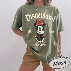 Może przedstawiać: Oliwkowa koszulka z grafiką Minnie Mouse, czerwoną kokardą i napisem "Disneyland est. 1955 California". Koszulka jest w stylu Comfort Colors Moss.