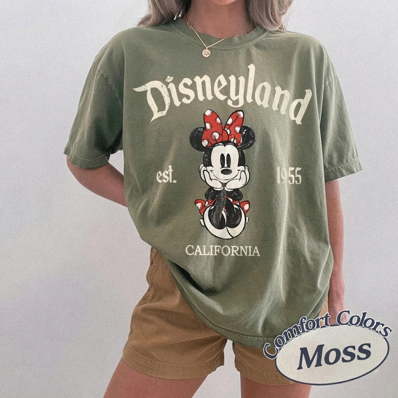 Może przedstawiać: Oliwkowa koszulka z grafiką Minnie Mouse, czerwoną kokardą i napisem "Disneyland est. 1955 California". Koszulka jest w stylu Comfort Colors Moss.