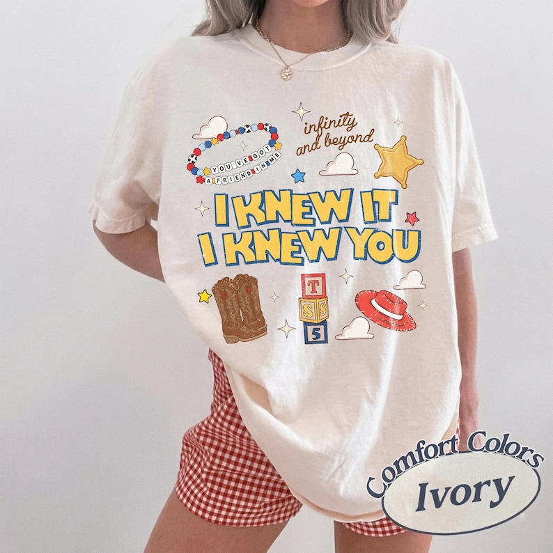 May include: An ivory short-sleeve t-shirt featuring a graphic print with the text I KNEW IT I KNEW YOU, infinity and beyond, and YOU'VE GOT A FRIEND IN ME. The design includes a beaded bracelet, a yellow star, cowboy boots, blocks, and a red hat.