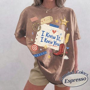 May include: An espresso-colored t-shirt featuring a central graphic of a torn piece of lined paper with the text "I Knew It, I Knew You" in blue. The design includes a red cowboy boot, a sheriff star, a wagon wheel, and various small stars.