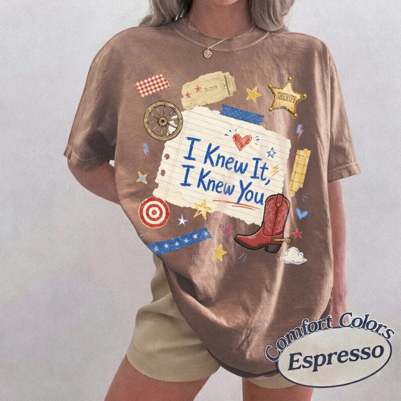 May include: An espresso-colored t-shirt featuring a central graphic of a torn piece of lined paper with the text "I Knew It, I Knew You" in blue. The design includes a red cowboy boot, a sheriff star, a wagon wheel, and various small stars.