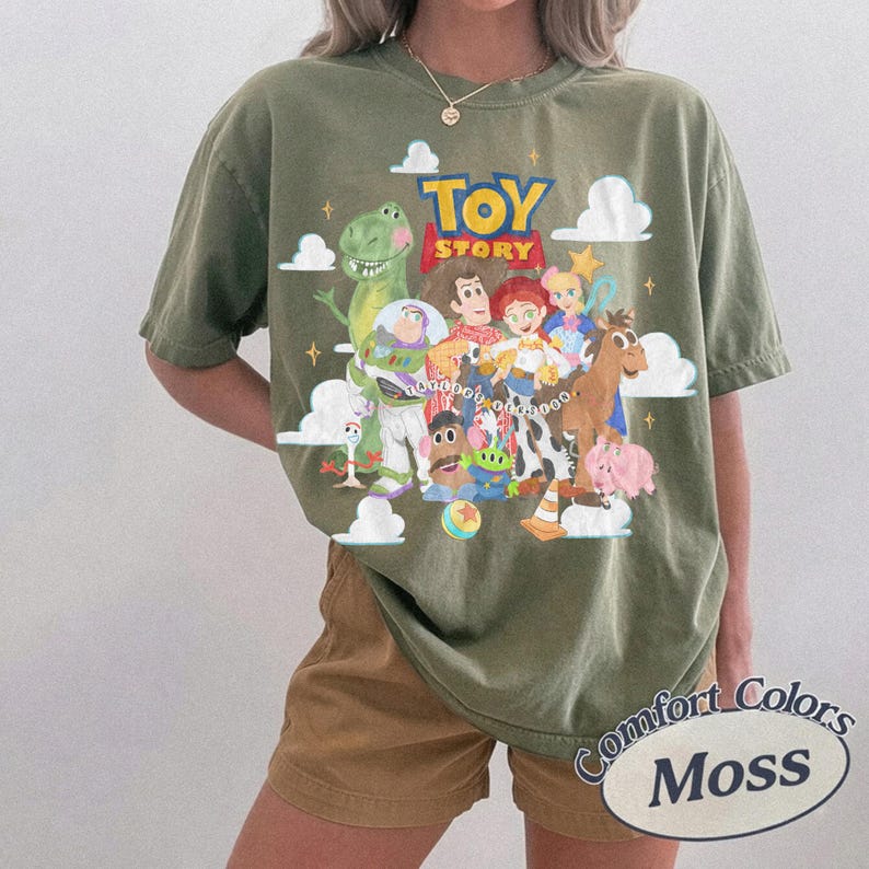 May include: An olive green short-sleeve t-shirt featuring a graphic print of Toy Story characters including Woody, Buzz Lightyear, Jessie, Rex, and others. The design includes white clouds, yellow stars, and the Toy Story logo in red and yellow text.