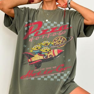 Pu&ograve; includere: T-shirt verde oliva con grafica di alieni, razzo e pianeta pizza. Il testo "Pizza H-O-T-T-O-Go" e "You Can Take Me H-O-T-T-O-Go" &egrave; stampato in rosso. Il design ha uno stile retr&ograve; e cartoonesco.