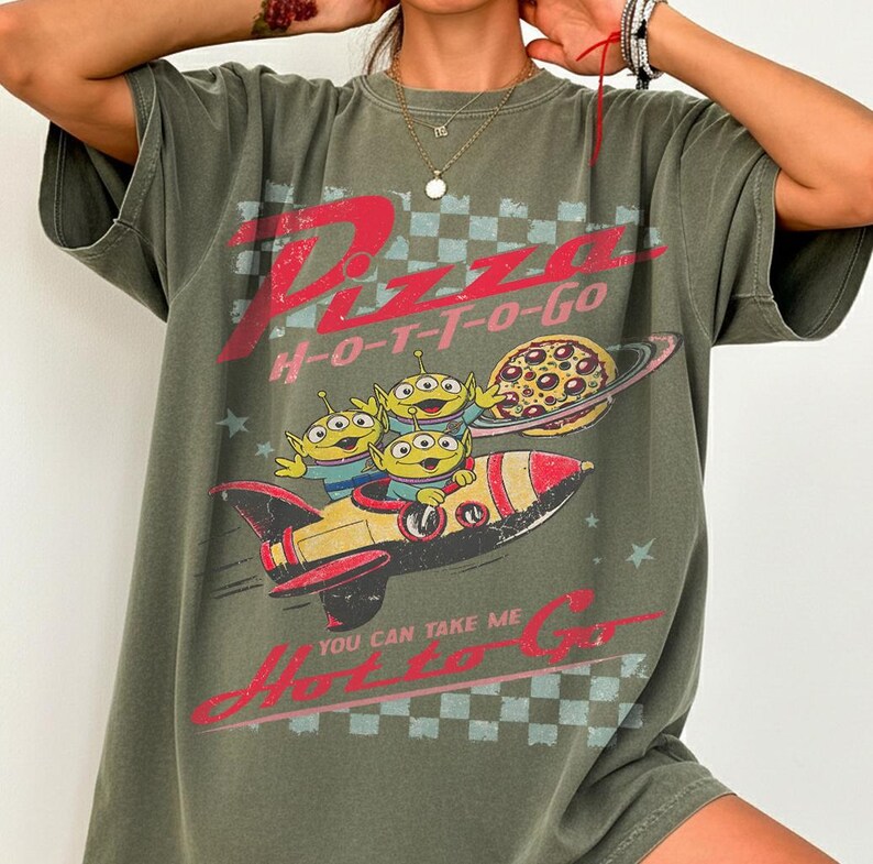 Pu&ograve; includere: T-shirt verde oliva con grafica di alieni, razzo e pianeta pizza. Il testo "Pizza H-O-T-T-O-Go" e "You Can Take Me H-O-T-T-O-Go" &egrave; stampato in rosso. Il design ha uno stile retr&ograve; e cartoonesco.