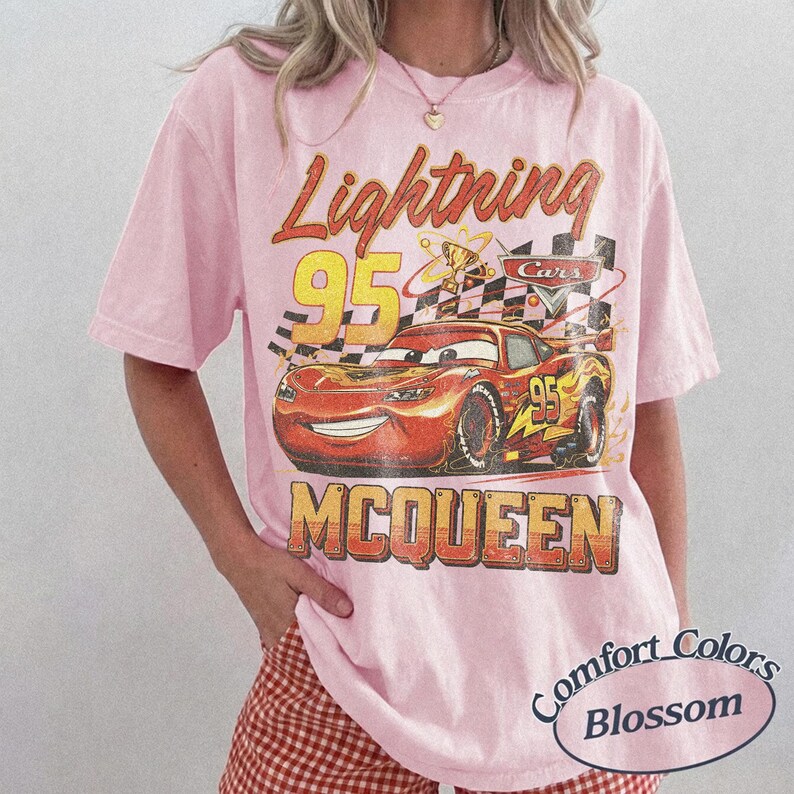 May include: Pink t-shirt featuring a graphic of Lightning McQueen from the Cars movie. The design includes the text "Lightning 95 McQueen" in a vintage style. The shirt is a Comfort Colors Blossom.