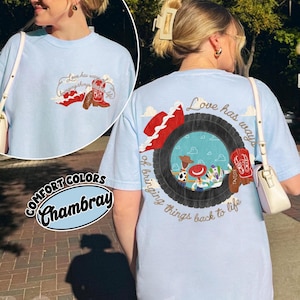 May include: A light blue t-shirt featuring a graphic on the back and front. The back design shows a black tyre framing a scene with cartoon characters, a red cowboy hat, and red boots, with the text Love has ways of bringing things back to life.