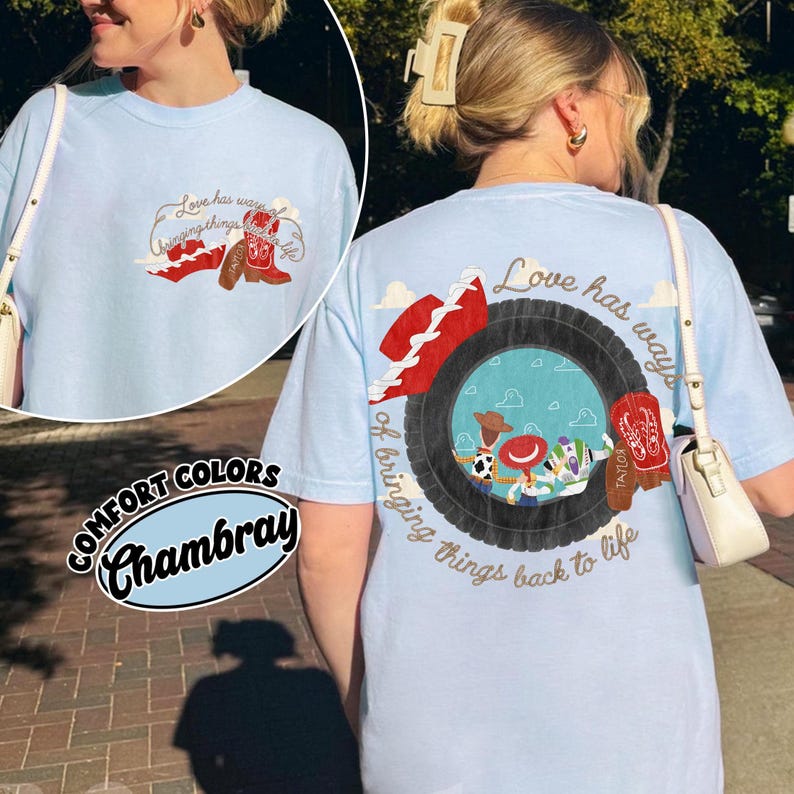 May include: A light blue t-shirt featuring a graphic on the back and front. The back design shows a black tyre framing a scene with cartoon characters, a red cowboy hat, and red boots, with the text Love has ways of bringing things back to life.