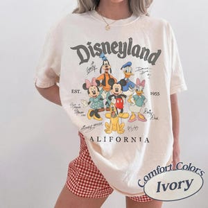 K&ouml;nnte beinhalten: Ein elfenbeinfarbenes Kurzarm-T-Shirt mit einer Grafik von Disney-Figuren wie Mickey Maus, Minnie Maus, Donald Duck, Daisy Duck, Goofy und Pluto. Der Text lautet Disneyland, Est. 1955 und California, erg&auml;nzt durch die Unterschriften der Figuren.