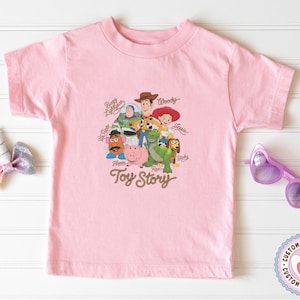 May include: A light pink short-sleeve t-shirt featuring a graphic print of Toy Story characters including Woody, Buzz Lightyear, Jessie, Mr. Potato Head, Hamm, Rex, and Slinky Dog, with the text Toy Story and individual character names printed.