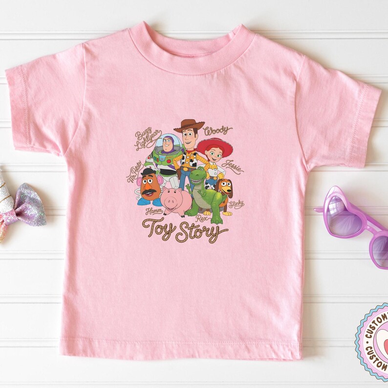 May include: A light pink short-sleeve t-shirt featuring a graphic print of Toy Story characters including Woody, Buzz Lightyear, Jessie, Mr. Potato Head, Hamm, Rex, and Slinky Dog, with the text Toy Story and individual character names printed.