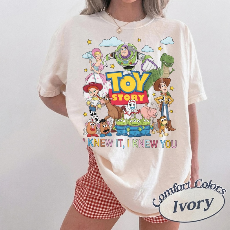 May include: An ivory short-sleeve t-shirt featuring a colorful graphic of Toy Story characters including Buzz Lightyear, Woody, Jessie, and others. The text "TOY STORY" is printed in the center, with "I KNEW IT, I KNEW YOU" written below in colorful letters.