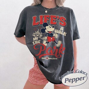 May include: Dark grey t-shirt with Mickey Mouse graphic and the text "Life's a Walk in the Park" in red and white. The shirt also includes "Walt Disney World" and a castle graphic. The shirt is a Comfort Colors brand.