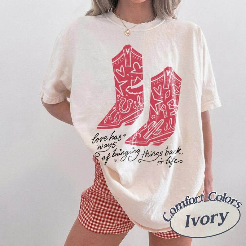 May include: An ivory short-sleeve t-shirt featuring a red graphic of two cowboy boots decorated with hearts, stars, and the number 13. Below the boots, the text reads, love has ways of bringing things back to life, in a black cursive script.