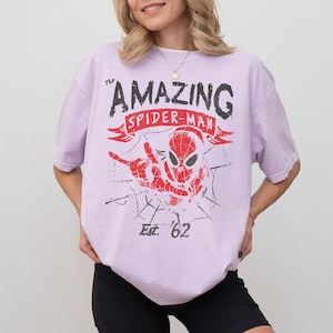 Pode incluir: Camiseta lil&aacute;s clara com um gr&aacute;fico desgastado do Homem-Aranha. O design inclui o texto "THE AMAZING SPIDER-MAN" acima de uma faixa vermelha e "Est. '62" abaixo da imagem do Homem-Aranha.