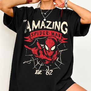 Pode incluir: Camiseta preta com um gr&aacute;fico vermelho e branco do Homem-Aranha. O design inclui as palavras "The Amazing Spider-Man" e "Est. '62". O super-her&oacute;i &eacute; retratado em uma pose din&acirc;mica com um fundo de teia de aranha.