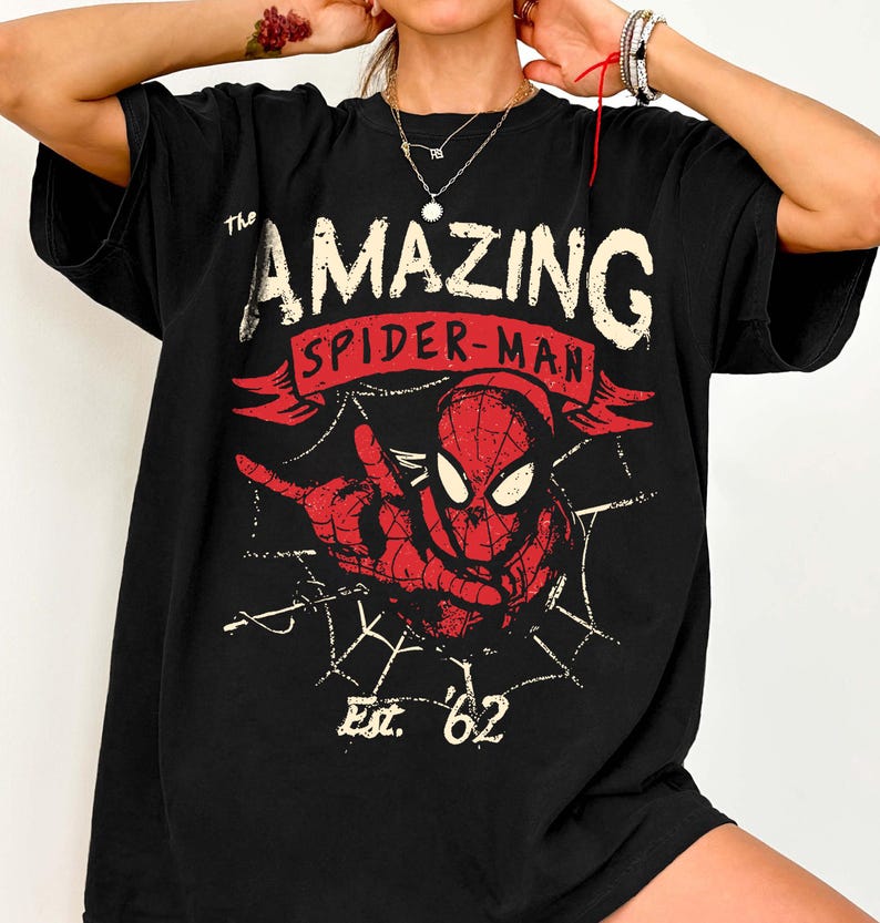 Pode incluir: Camiseta preta com um gr&aacute;fico vermelho e branco do Homem-Aranha. O design inclui as palavras "The Amazing Spider-Man" e "Est. '62". O super-her&oacute;i &eacute; retratado em uma pose din&acirc;mica com um fundo de teia de aranha.