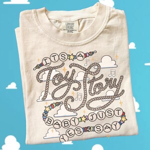 May include: A cream-colored short-sleeve t-shirt featuring a graphic design with the text 'ITS A Toy Story BABY JUST YES SAY'. The lettering is styled like rope, surrounded by colorful beads, stars, musical notes, and cloud illustrations.